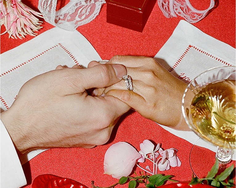 Close-up of two people holding hands at a red table, showing an engagement ring and wedding band.