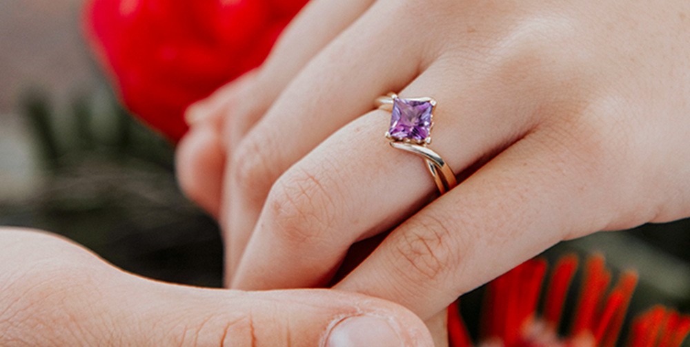 Hand wearing purple gemstone engagement ring against floral background.