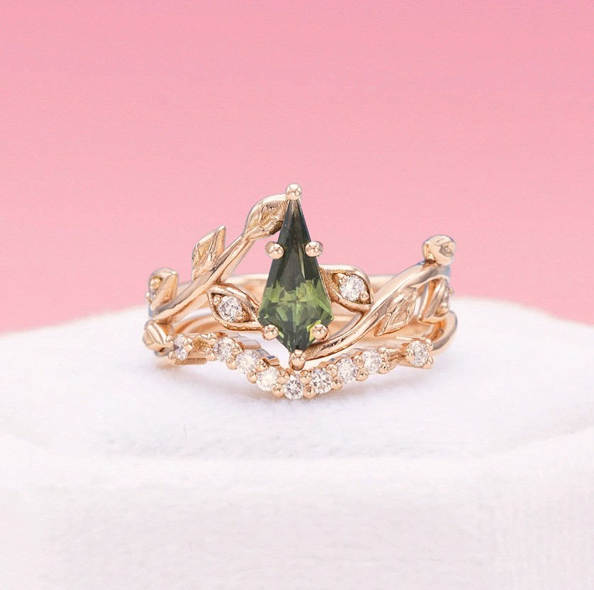 Nature-inspired gold engagement ring featuring green pear-shaped gemstone and diamond leaf detailing, shown on pink backdrop.